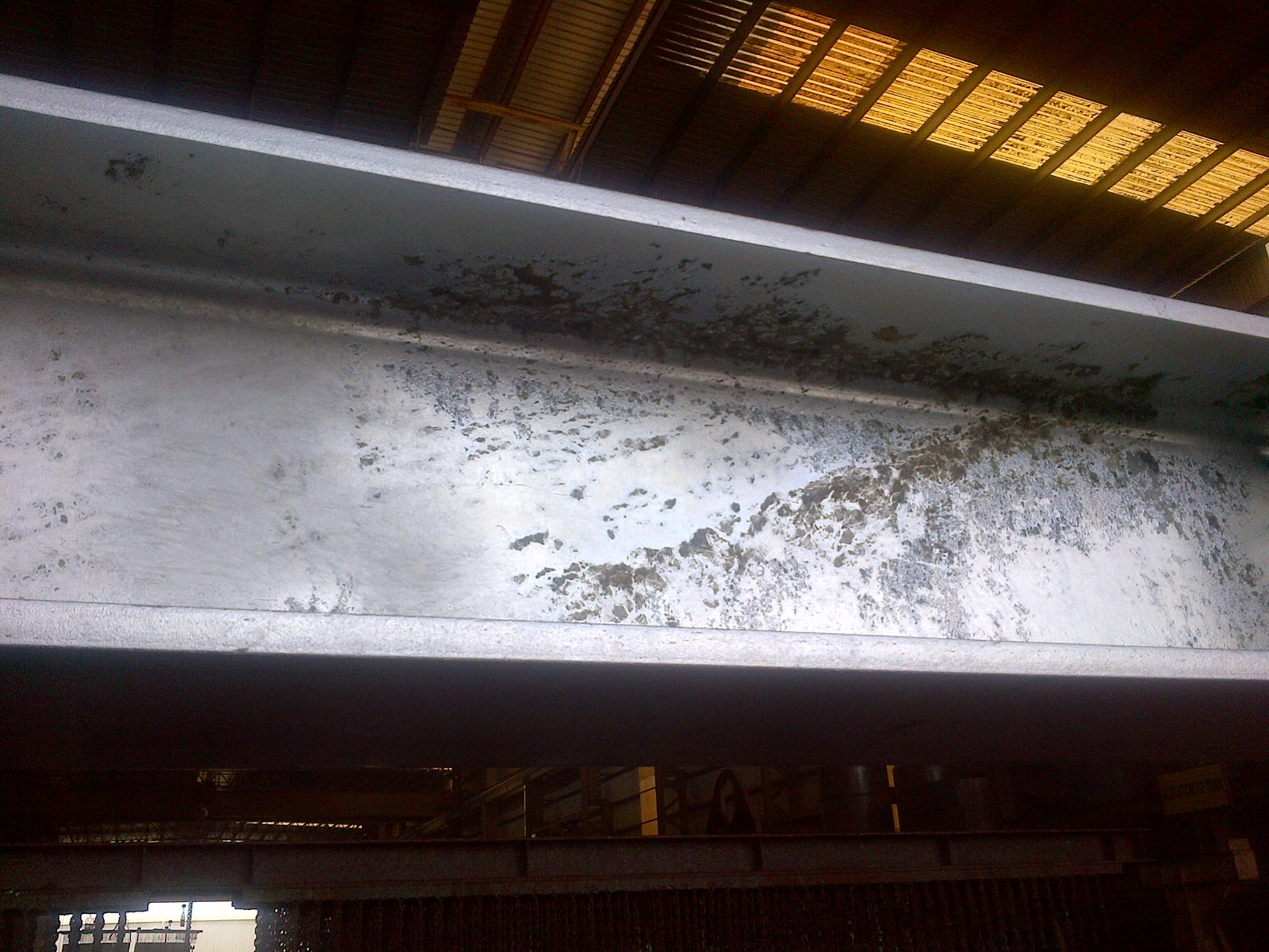 Galvanized beam photographed from below: a broad chalky grey-white ash deposit spreads across the web with soft feathered edges, clearly lighter than the sound zinc beside it, and the same deposit continues as a dark band under the top flange where it falls into shadow