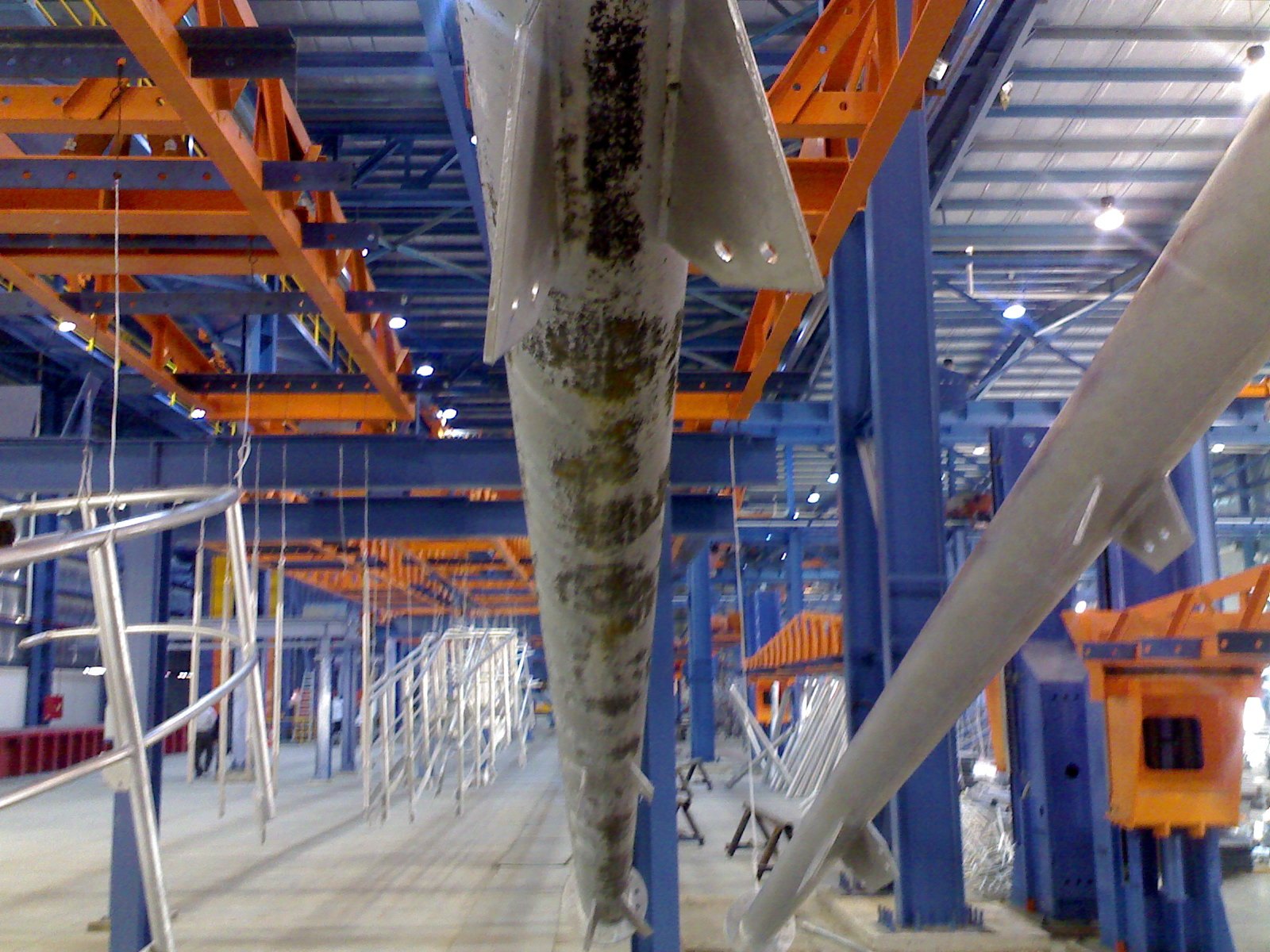 A tapered galvanized pole hanging on a rack in the plant, seen from below, with dark speckled patches streaking down one side of the otherwise pale coating