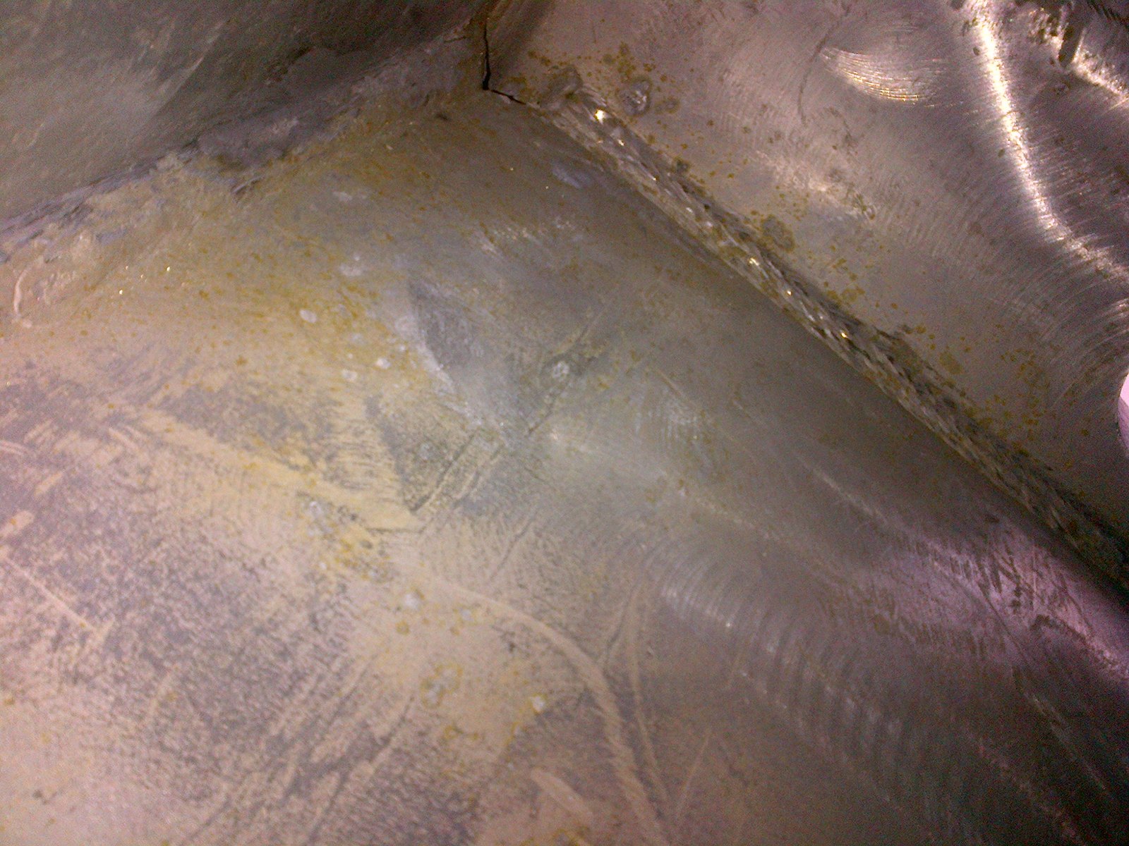 Close-up of a galvanized fillet weld corner: a broad translucent yellow-ochre flux film lies across the flange face with fine white crystalline speckles through it, while the weld bead itself is fully galvanized with its ripples clearly visible under sound zinc
