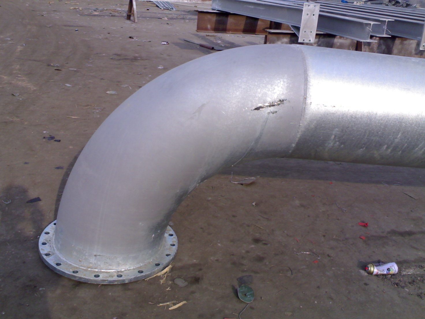 Large galvanized pipe bend with a flange: the elbow shows a dull matte grey coating while the adjoining straight pipe shows bright spangle, in the same assembly
