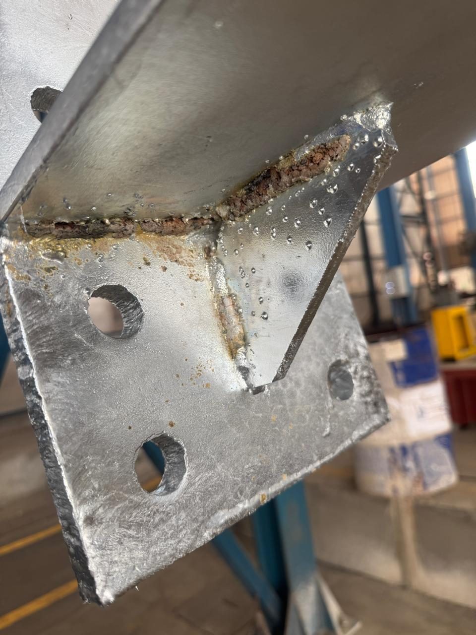 Galvanized gusset and base-plate connection: a brown crusty slag ridge runs along the weld toes with no zinc on it, rust stain bleeds onto the coating below, and round weld spatter nodules are scattered across the gusset face