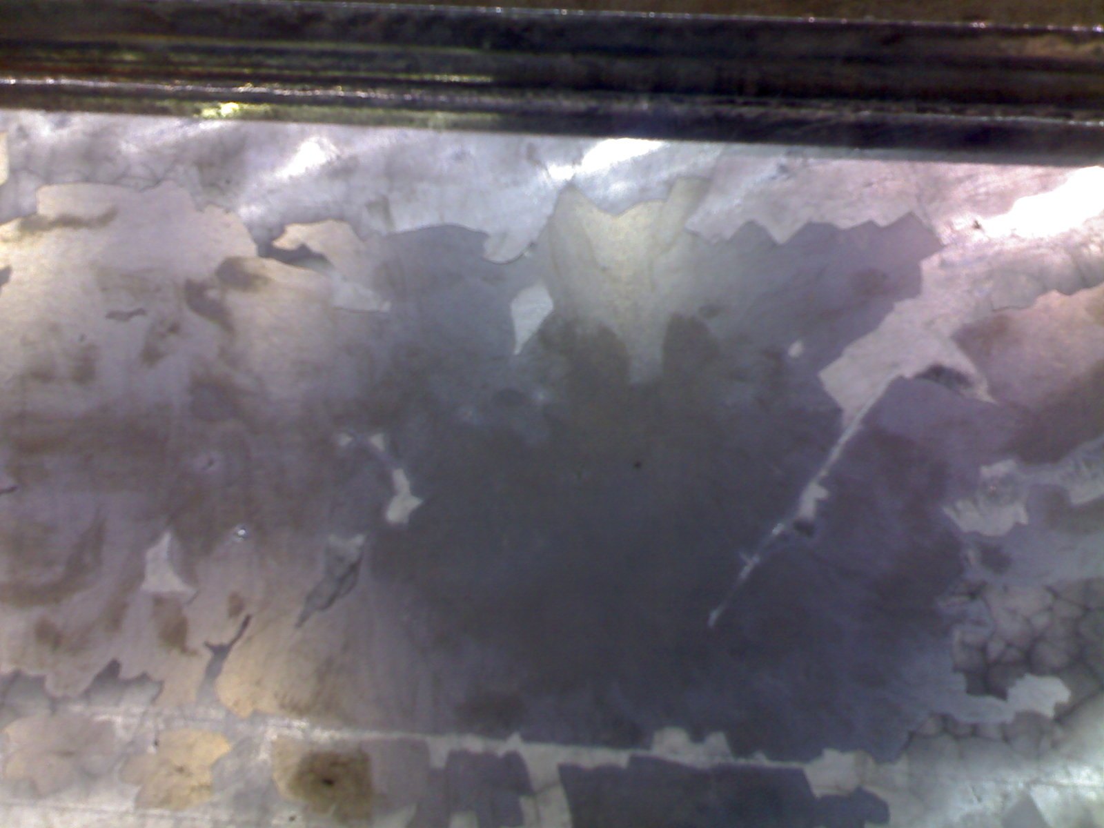 Galvanized surface where the bright free zinc has flaked away across the centre, exposing the darker smooth zinc-iron alloy layer beneath, with the remaining zinc surviving around the edges as chalky islands with jagged boundaries